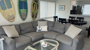 A living room with a gray sectional sofa, a glass coffee table, and two surfboards on the wall. - 3 bedroom, 2.5 bath unit - Ocean, bay and pool all in a day! (Lewes)