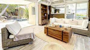A spacious living area with a wicker sofa, a wooden coffee table, and a large window offering a view of a pool and outdoor area. - Villa Cazou, vue mer, piscine privée, Wifi 100Mbps (Oyster Pond)