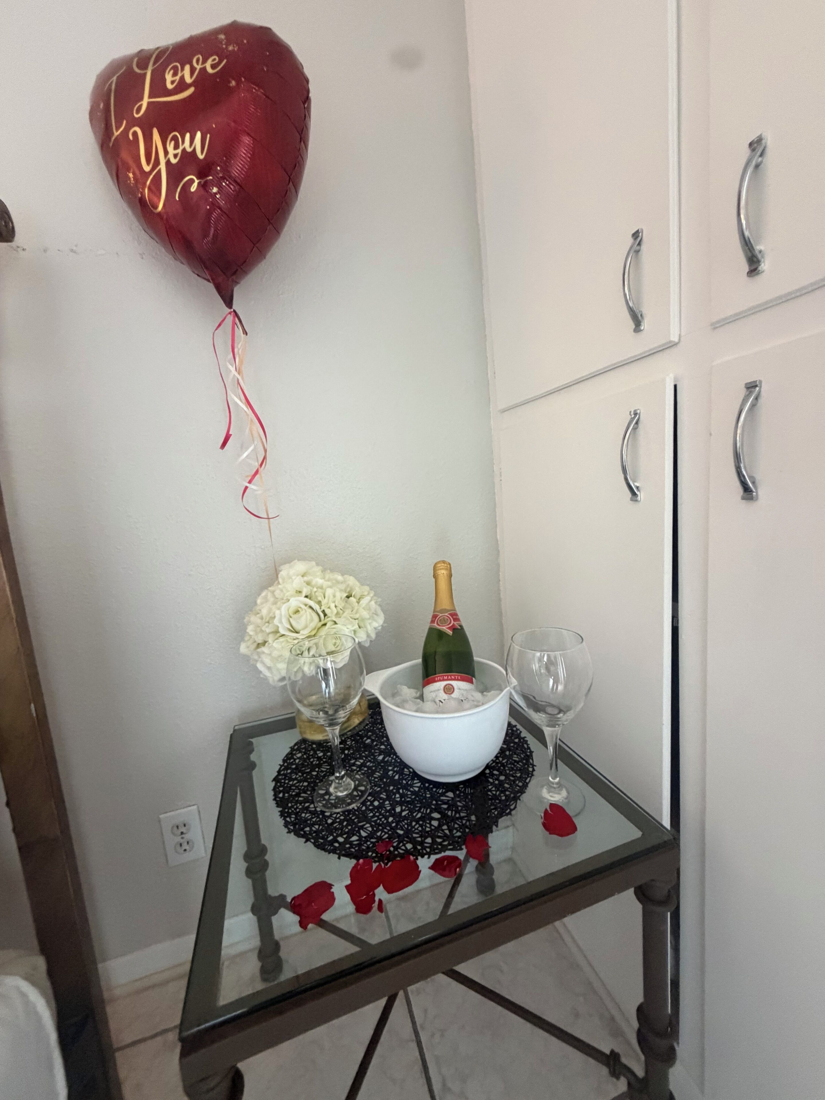 A table with a bottle of champagne, two glasses, a bowl of flowers, and a heart-shaped balloon with 'I Love You' written on it.