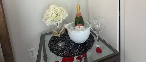 A table with a bottle of champagne, two glasses, a bowl of flowers, and a heart-shaped balloon with 'I Love You' written on it.