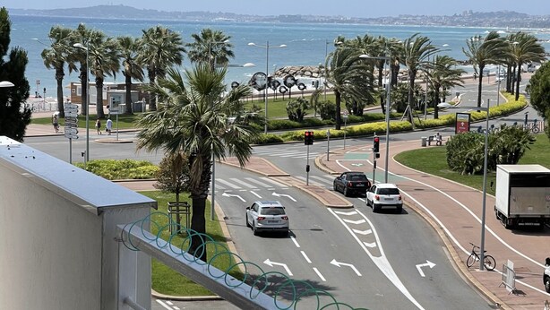 A balcony with a table displaying a menu, overlooking a busy street with palm trees and a view of the sea. - Appartement Climatisé Bord de mer Refait a Neuf (Cagnes-sur-Mer)