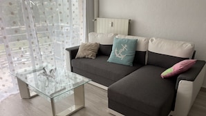 A modern living room with a glass coffee table, a grey sofa with multiple cushions, and a view through sheer curtains. - Appartement Climatisé Bord de mer Refait a Neuf (Cagnes-sur-Mer)