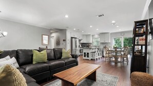Een moderne woonkamer met een leren zwarte bank, een houten salontafel en een eethoek met een witte tafel en stoelen. In de achtergrond is een keuken te zien met witte kasten en roestvrijstalen apparatuur.