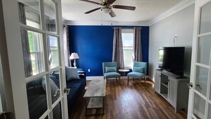 Una sala de estar con pared azul, piso de madera, ventilador de techo, sofá, dos sillones, una mesa de centro y un mueble para televisión.
