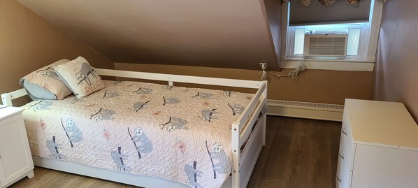 A bedroom with a bed, pillows, a nightstand, a dresser, and a window with an air conditioning unit. - Casinos, beaches, and history (Norwich)