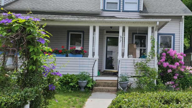 Una casa de dos pisos con un porche, plantas en macetas y una valla de madera.