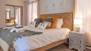 Una cama bien hecha con varias almohadas y una manta, una mesita de noche con una lámpara y un espejo en la pared.