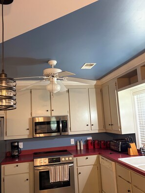 A kitchen with white cabinets, a stainless steel microwave, a gas stove, a ceiling fan, and a window with blinds.