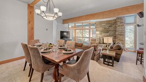 A dining area with a wooden table set for four, surrounded by cushioned chairs. A stone wall and large windows provide natural light and views. - Powder Run 12C 100 yards to Lift Unbeatable (Park City)