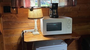 A compact kitchenette with a microwave, coffee maker, and a small refrigerator. A window with curtains allows natural light and a view of the outdoors. - Rustic Bee Cabin - camping (Hardin)
