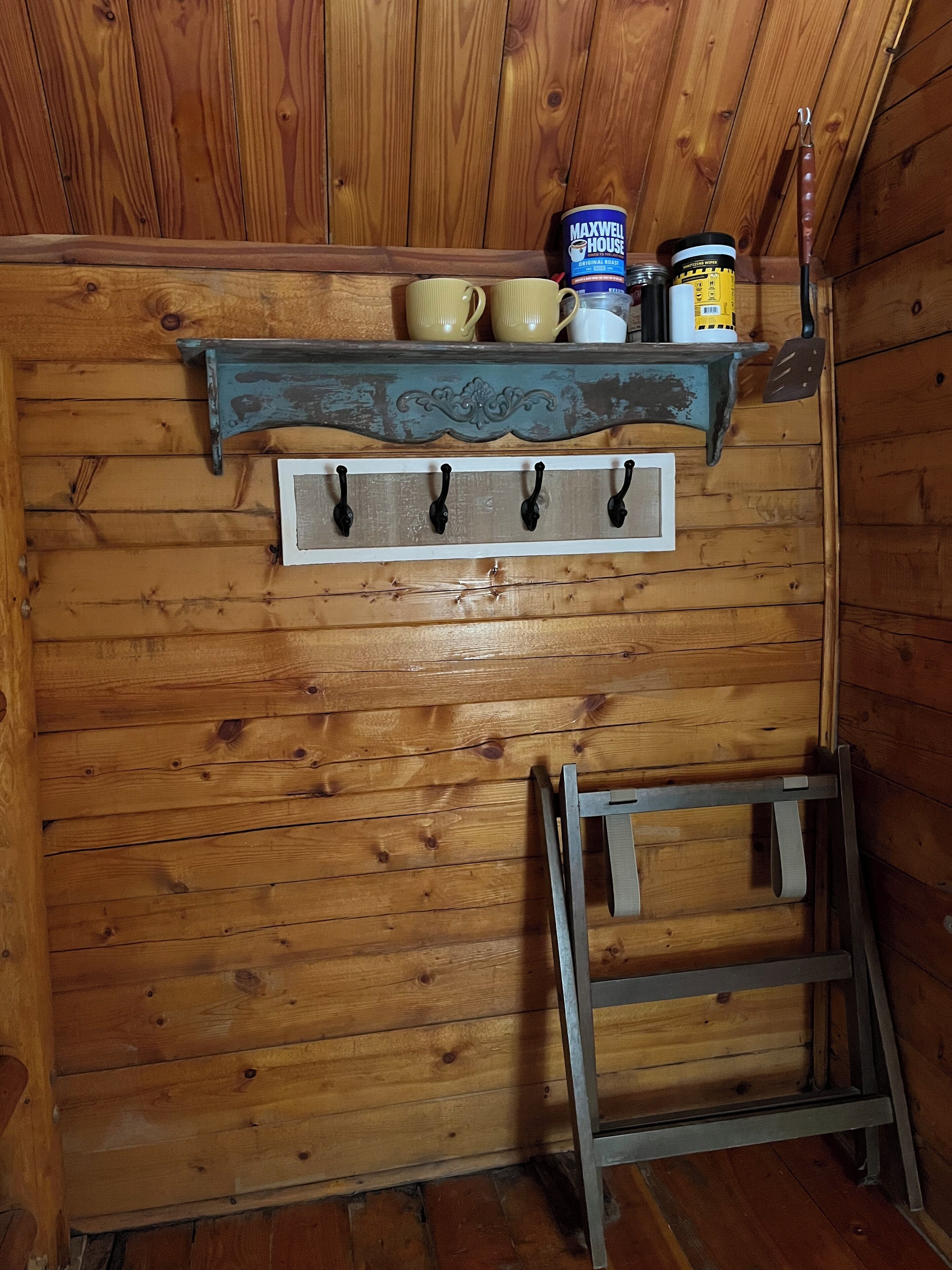 A wooden interior with a shelf holding mugs, a can of Maxwell House coffee, and a ladder.