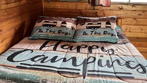 A wooden cabin room with a bed featuring a 'Happy Campers' themed bedspread and matching pillows. - Rustic Bee Cabin - camping (Hardin)