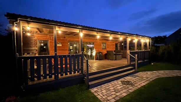 Een houten chalet met een overdekte veranda, zitplaatsen buiten en een met baksteen bestraat pad dat naar de ingang leidt.