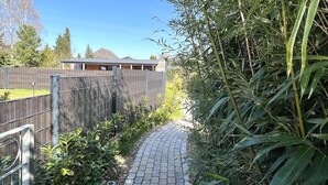 Een geplaveid pad, omzoomd door weelderige begroeiing en een metalen hek, leidt naar een gebouw met een schuin dak.