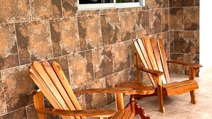 Two wooden Adirondack chairs with a small table, positioned on a tiled floor against a wall with large, square tiles.