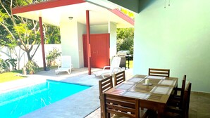 A poolside dining area with a table and chairs, a barbecue grill, and a covered seating area with chairs.