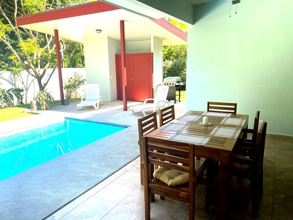 Un área de comedor junto a la piscina con mesa y sillas, una parrilla y un espacio de estar cubierto con sillas.