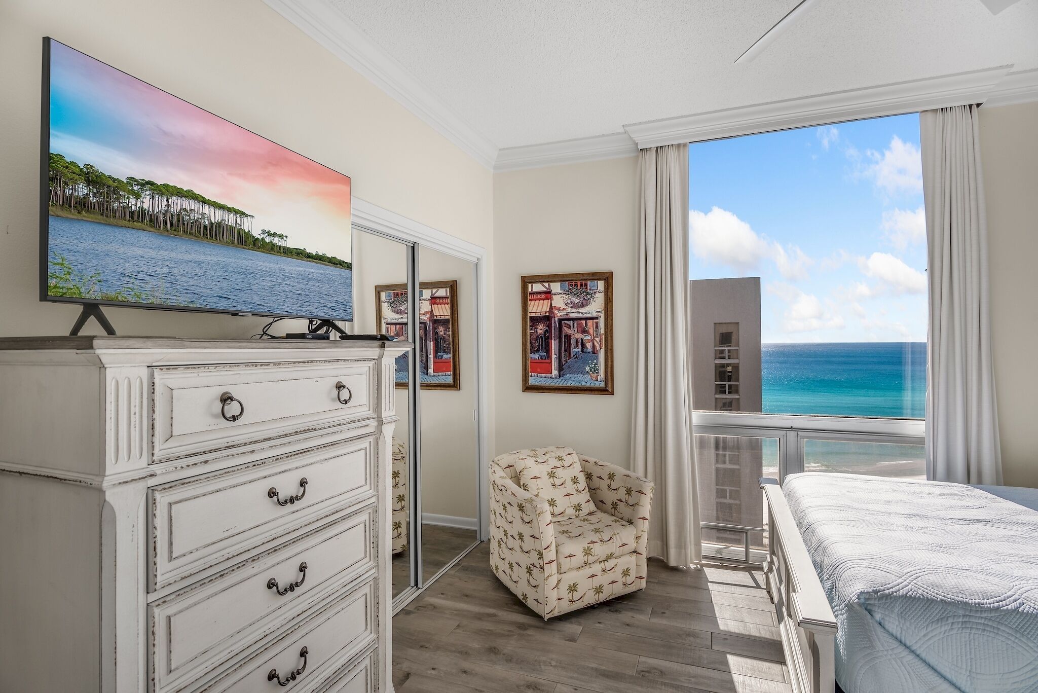 A bedroom with a large TV, a chair, a bed, and a balcony with a view of the ocean.