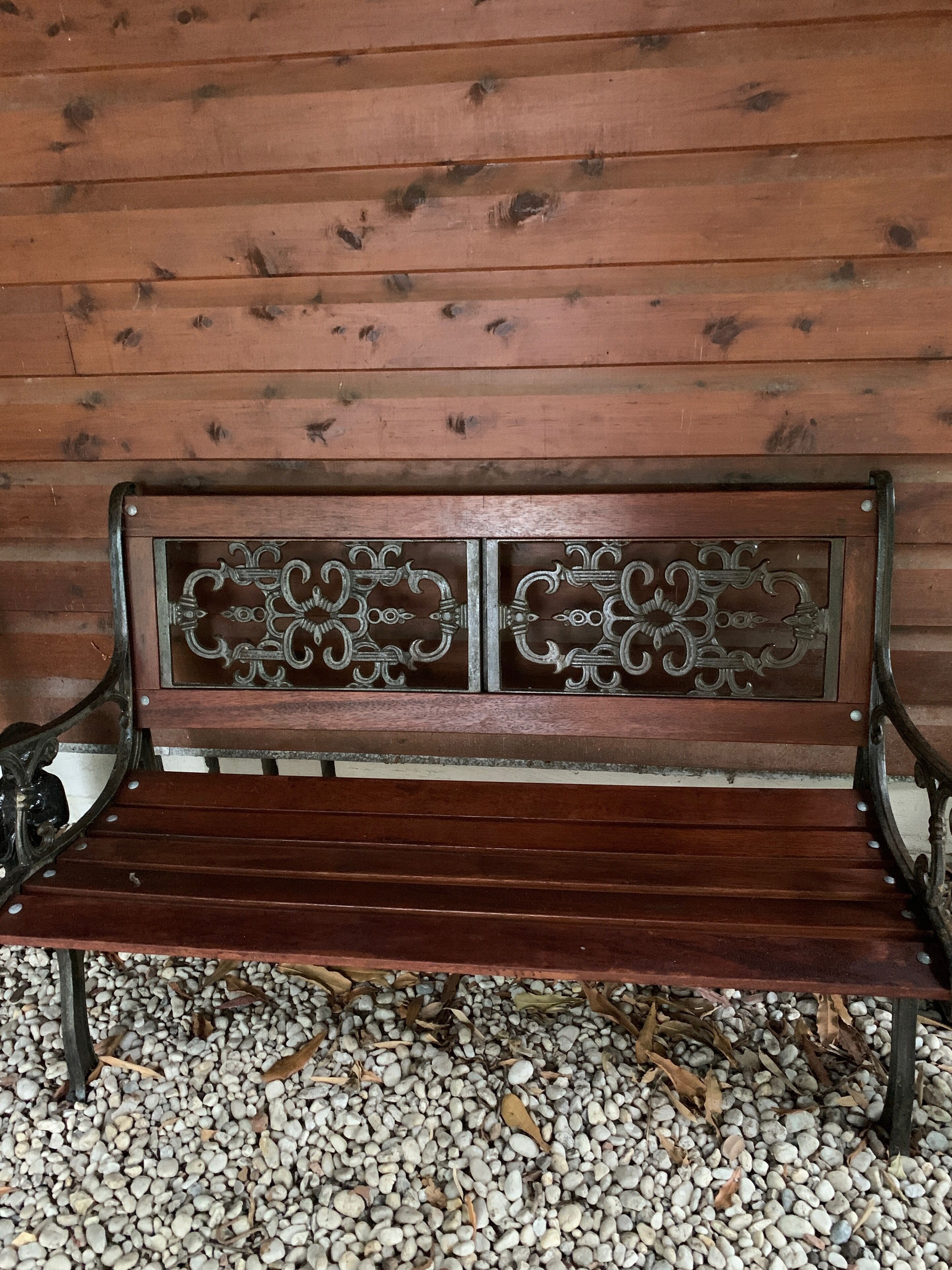 A wooden bench with metal armrests and decorative panels, set on a gravel surface against a wooden wall.