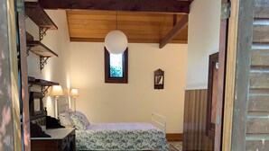Ein Zimmer mit Holzboden, einem Bett mit Blumenmuster am Überwurf, einem Fenster mit Blick auf Grünflächen und einem kleinen Tisch mit Lampe.