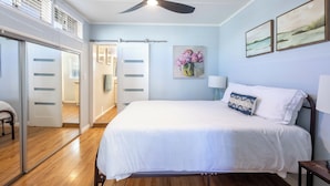 A bedroom with a bed, a ceiling fan, a painting, and a door leading to a bathroom. - Across quiet street from sandy beach. (Sunset Beach)