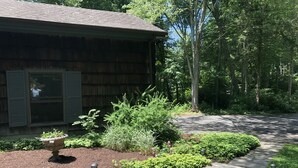 Une cabane en bois, avec un chemin de pierre menant à l’entrée, entourée d’une végétation luxuriante et d’arbres.