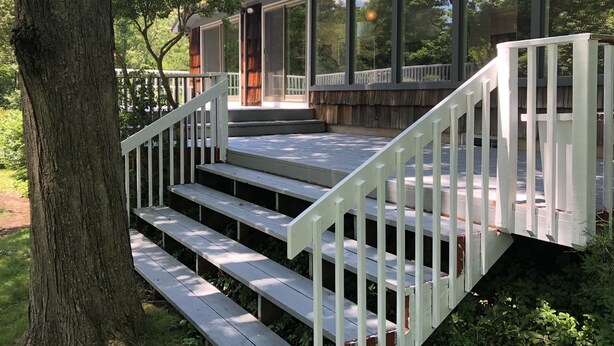 Une terrasse en bois avec des balustrades blanches, qui mène à un porche couvert doté de grandes fenêtres.