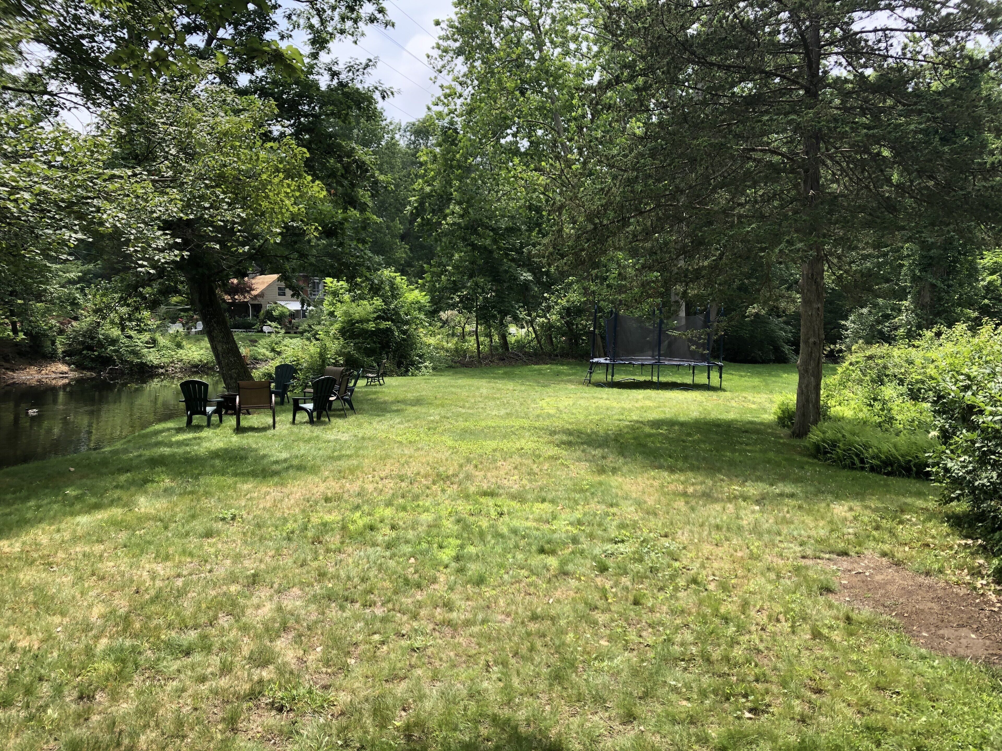 A grassy backyard with a trampoline, chairs, and a pond.