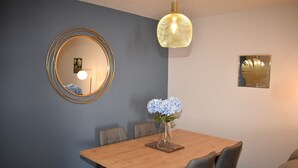 Een eetgedeelte met een houten tafel, grijze stoelen, een vaas met blauwe bloemen, een ronde spiegel en een hangende lamp.