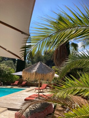 Uma área na beira da piscina com espreguiçadeiras, um guarda-sol de palha e palmeiras.