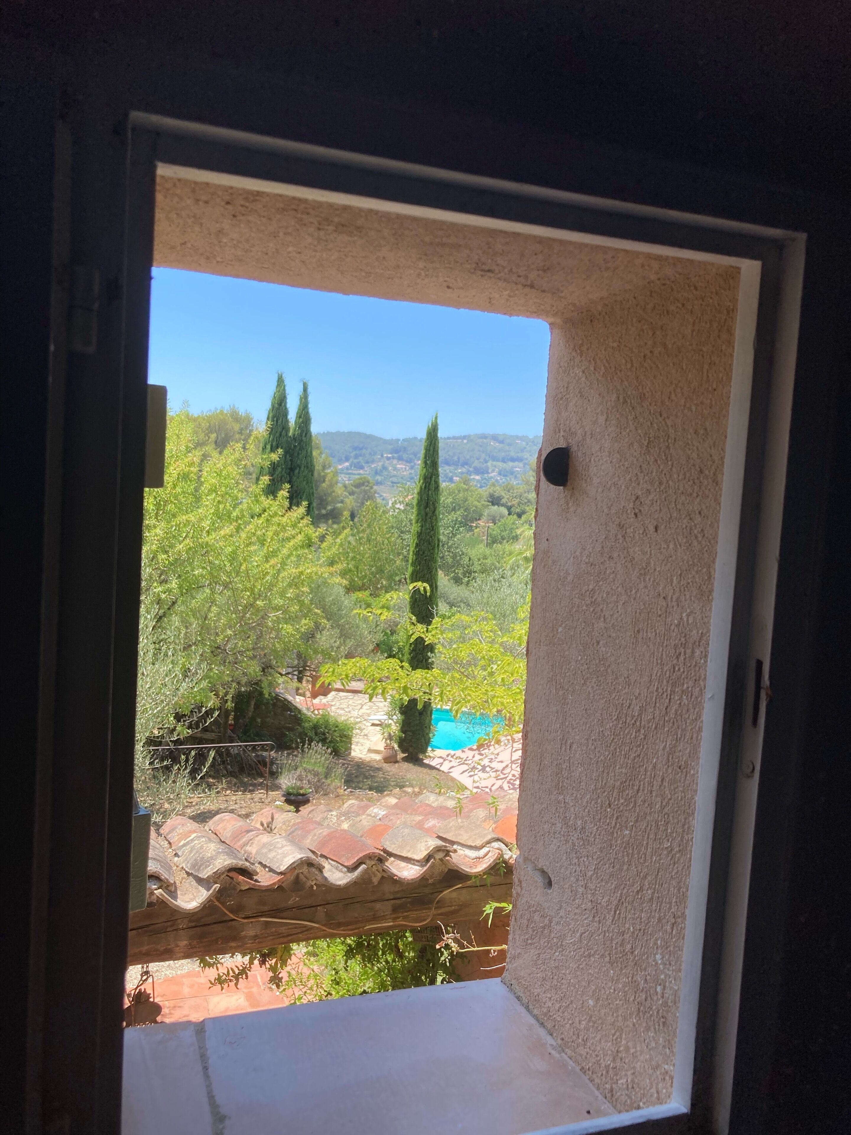 Vista a través de una puerta abierta que da a un cuarto con piso de baldosas y una ventana con vista a un jardín con árboles y una piscina.