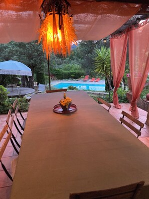 Uma mesa de jantar comprida com cadeiras, um arranjo decorativo, um guarda-sol e uma piscina ao fundo.