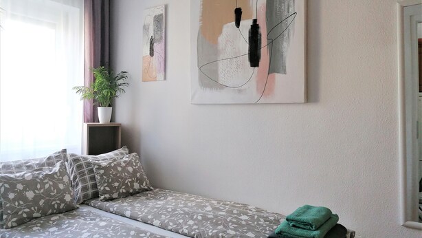 Ein modernes Schlafzimmer mit Schlafsofa, Topfpflanze, abstrakten Wandbildern und einem Stapel Handtücher.