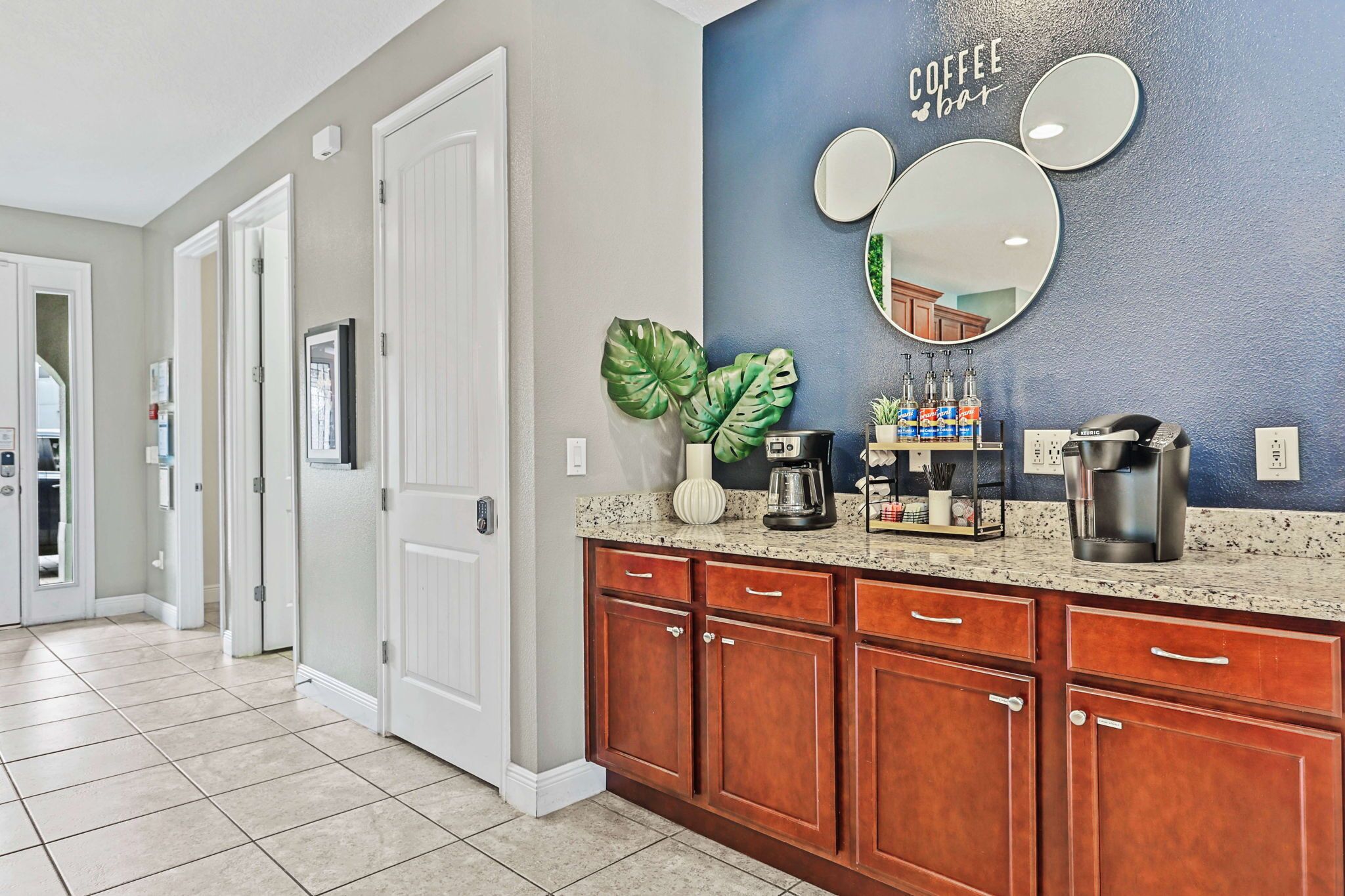 A kitchen with wooden cabinets, a granite countertop, a coffee station, and a wall with a coffee-themed mirror and artwork.