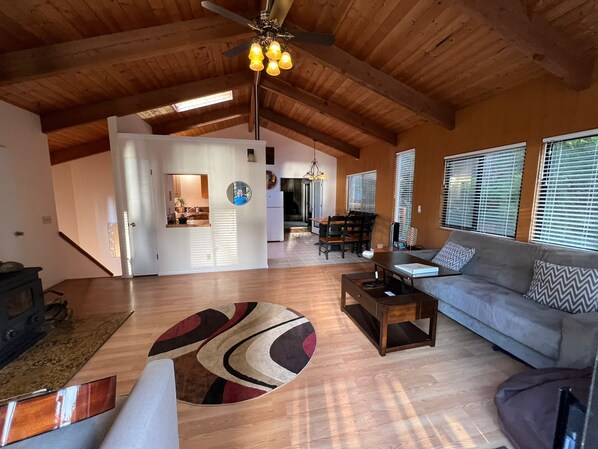 A spacious living room with a high ceiling, wooden beams, a dining area, and a kitchen visible through an open door.