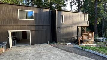 A two-story wooden cabin with a garage entrance, a small deck, and a surrounding forest.