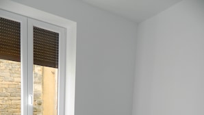 Ein kleines Zimmer mit einem Einzelbett, einem Holz-Kopfteil, einem Fenster mit Jalousien und einer dahinter liegenden Steinwand.
