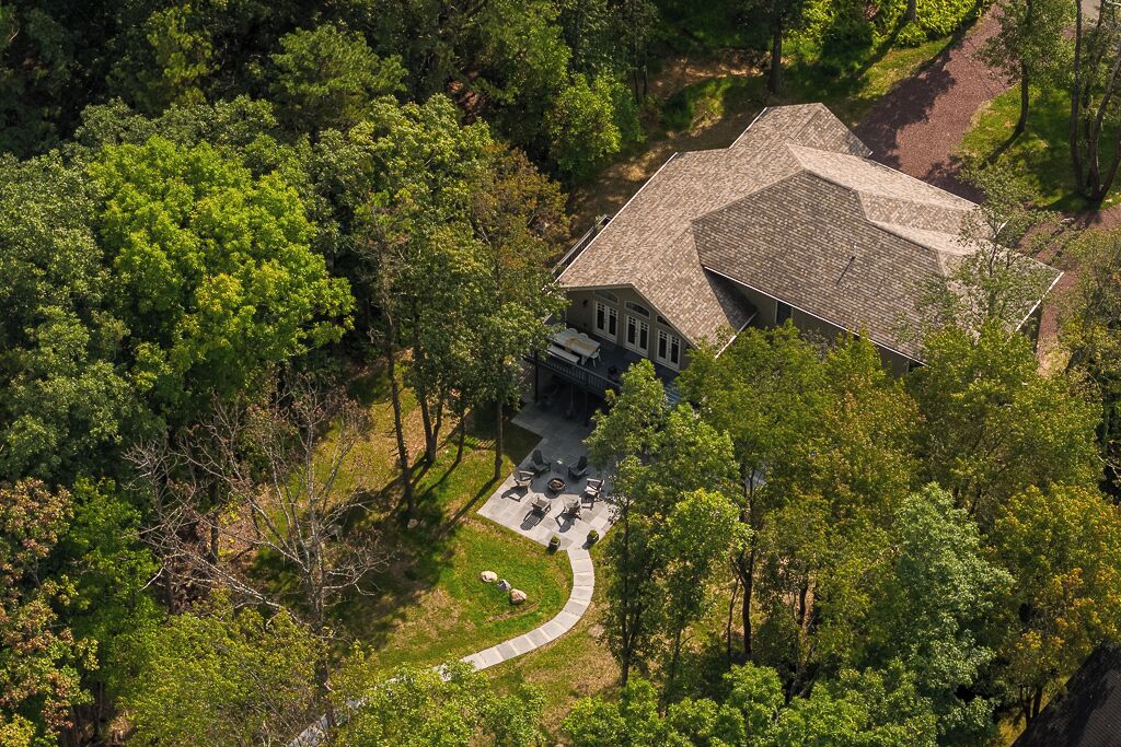 An aerial view of a house surrounded by dense trees, with a paved driveway leading to a patio area.