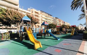 Children can play on the slides and climbing structures in this playground area, surrounded by a green rubberized surface and a paved walkway.