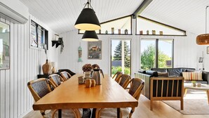 Ett rymligt matområde med ett långt träbord och flera stolar, stora fönster med utsikt över grönområden och en mysig sittgrupp med en soffa och kuddar.