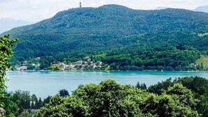A lakeside view with a mountain in the background, a small settlement with buildings near the water, and a tower on the mountain peak. - modernes Ferienhaus mit Garten (Techelsberg)