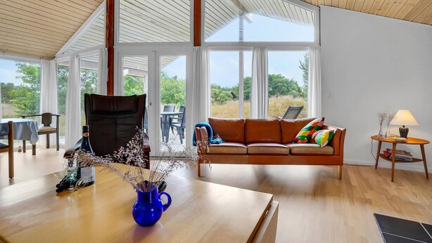 A spacious living room with a wooden ceiling, large windows, a brown leather sofa, a blue vase with dried flowers, and a wooden table. - 3 bedroom lovely home in Fanø (Fanø)
