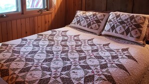 Ein Schlafzimmer einer Holzhütte mit einer gesteppten Bettdecke, zwei Kissen, einem Nachttisch und einem Fenster mit Vorhängen.