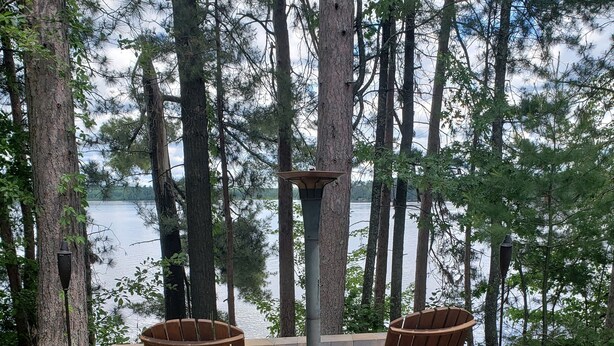 Eine Holzterrasse mit zwei Adirondack-Stühlen und einem Tisch, umgeben von Bäumen und mit Blick auf einen See.