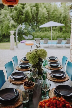 An outdoor dining area with a long table set for a meal, surrounded by chairs and a pool in the background. - Villa Leandra - Modern private villa near the lake and town (Gardone Riviera)