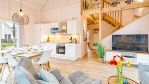 En rummelig opholdsstue med spisebord, behagelige siddepladser, et køkken med moderne hårde hvidevarer og en trappe, samt et stort fjernsyn.