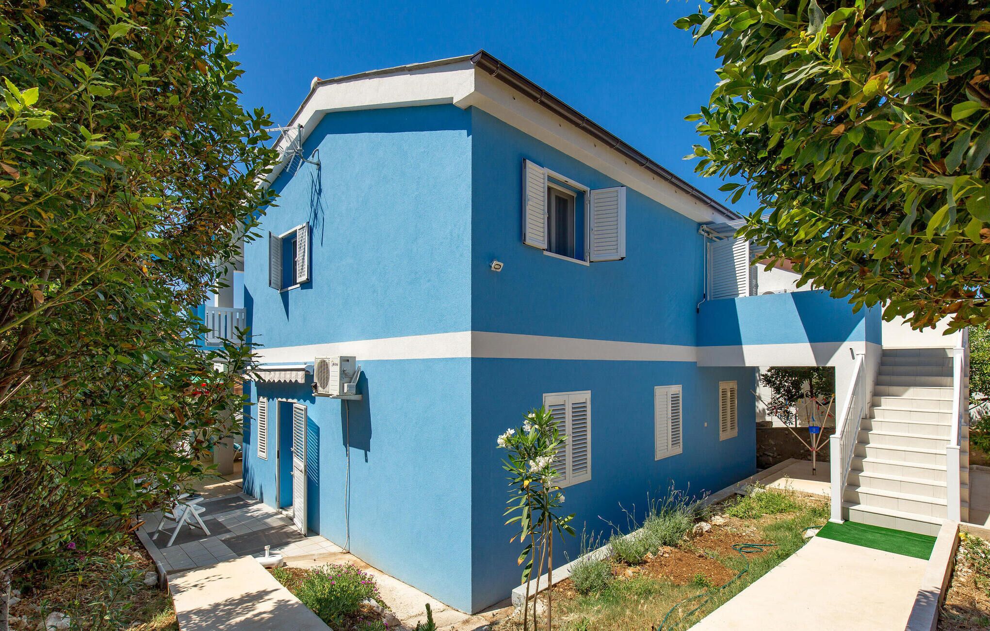 A two-story building with a blue exterior and white trim. There are multiple windows with shutters, and a small garden area with a pathway leading to the entrance.