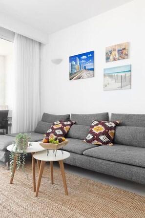 A modern living room with a grey sofa, two patterned throw pillows, a small round table with a bowl of fruit, and two wall-mounted paintings. - Panoramic Seaview Balcony Escape (Tel Aviv-Yafo)