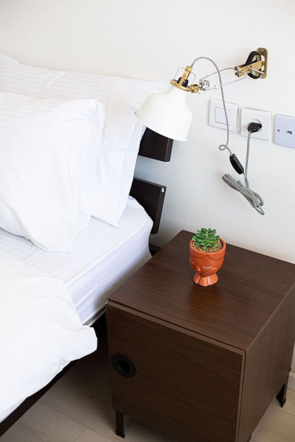 Un lit bien fait, avec du linge de lit blanc, une lampe de chevet, une petite plante en pot et un téléphone fixé au mur.