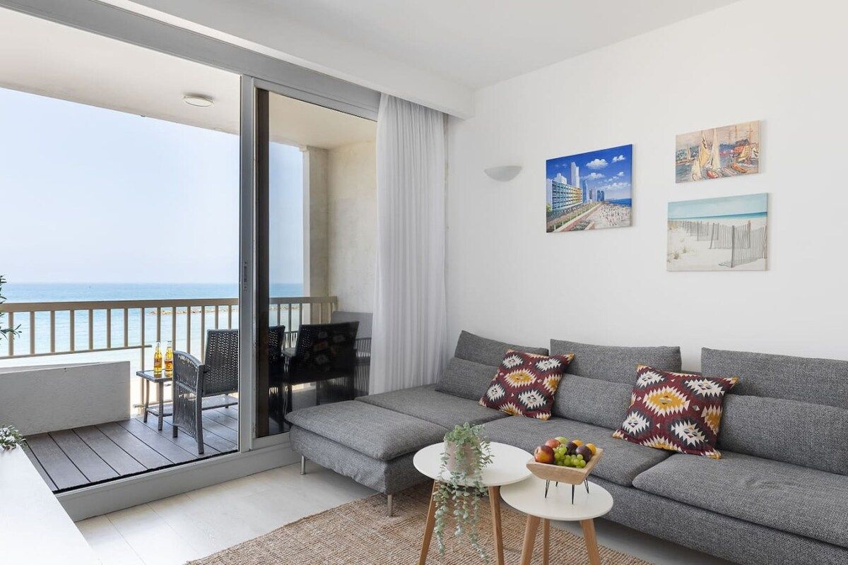 Un salon moderne avec un canapé gris, deux coussins à motifs, une petite table ronde sur laquelle est posé un panier de fruits, et une vue sur la mer par une porte-fenêtre coulissante.